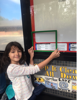 child holding frame in front of bus fares listed at their eye level